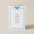 A white greeting card standing upright on a light beige surface, featuring a blue bow illustration at the top and placeholder text that reads 'ADD YOUR OWN TEXT HERE' inside a wavy blue border.