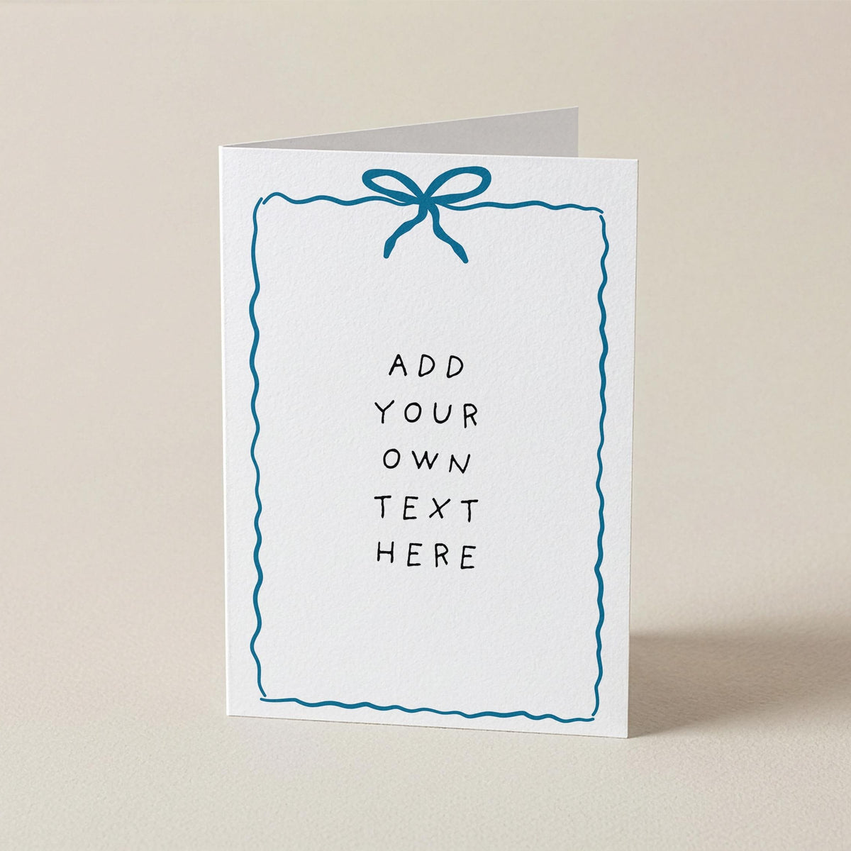 A white greeting card standing upright on a light beige surface, featuring a blue bow illustration at the top and placeholder text that reads 'ADD YOUR OWN TEXT HERE' inside a wavy blue border.