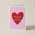 A folded greeting card standing upright on a light beige surface. The card has a pink background with a large red heart in the center and white text reading 'happy birthday to my SOUL SISTER'.