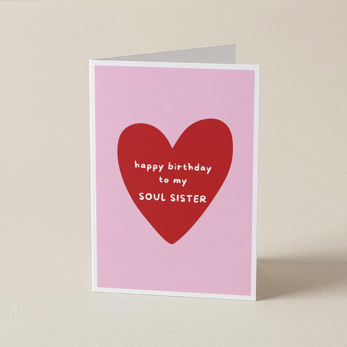 A folded greeting card standing upright on a light beige surface. The card has a pink background with a large red heart in the center and white text reading 'happy birthday to my SOUL SISTER'.