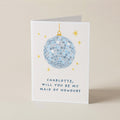 A white square greeting card standing upright on a beige surface, featuring a blue mirrored disco ball illustration with small gold stars and the text, 'CHARLOTTE, WILL YOU BE MY MAID OF HONOUR?'.