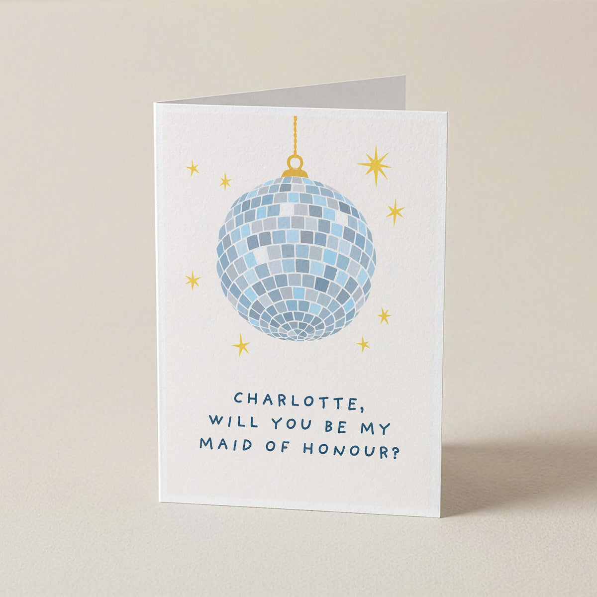 A white square greeting card standing upright on a beige surface, featuring a blue mirrored disco ball illustration with small gold stars and the text, 'CHARLOTTE, WILL YOU BE MY MAID OF HONOUR?'.