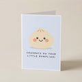 A folded greeting card standing upright on a light beige surface. The front of the card has a pale blue background with a cute cartoon dumpling face and the text 'CONGRATS ON YOUR LITTLE DUMBLING!'.