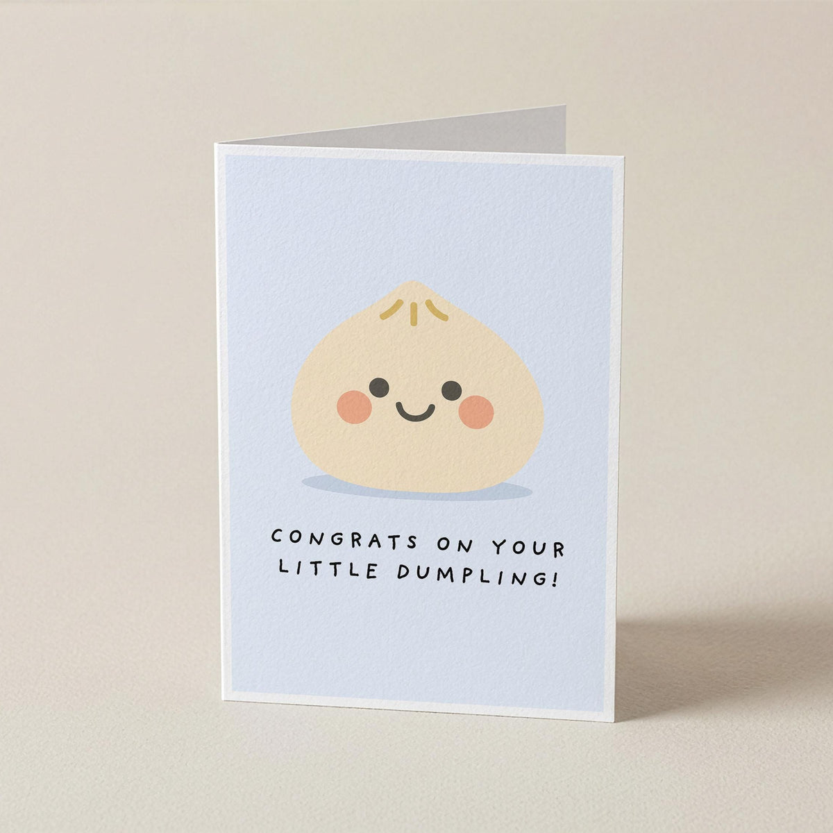 A folded greeting card standing upright on a light beige surface. The front of the card has a pale blue background with a cute cartoon dumpling face and the text 'CONGRATS ON YOUR LITTLE DUMBLING!'.