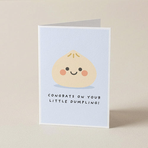 A folded greeting card standing upright on a light beige surface. The front of the card has a pale blue background with a cute cartoon dumpling face and the text 'CONGRATS ON YOUR LITTLE DUMBLING!'.