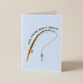 A folded greeting card standing upright on a beige surface. The card has a light blue background and features a fishing rod graphic with the text 'DAD, YOU'RE REELY GREAT!' above a small fish illustration.