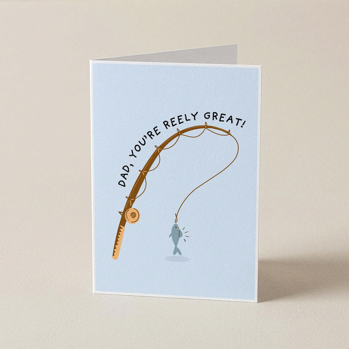 A folded greeting card standing upright on a beige surface. The card has a light blue background and features a fishing rod graphic with the text 'DAD, YOU'RE REELY GREAT!' above a small fish illustration.