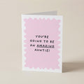 A folded greeting card standing upright on a light beige surface. The card has a white border with a scalloped light pink inner design and black text that reads, 'YOU'RE GOING TO BE AN AMAZING AUNTIE!'.