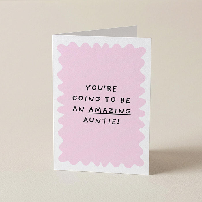 A folded greeting card standing upright on a light beige surface. The card has a white border with a scalloped light pink inner design and black text that reads, 'YOU'RE GOING TO BE AN AMAZING AUNTIE!'.