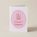 A folded greeting card standing upright on a light beige surface. The card is white with a pink scalloped oval graphic featuring a disco ball illustration and the text 'ELEANOR IS THIRTY!'.