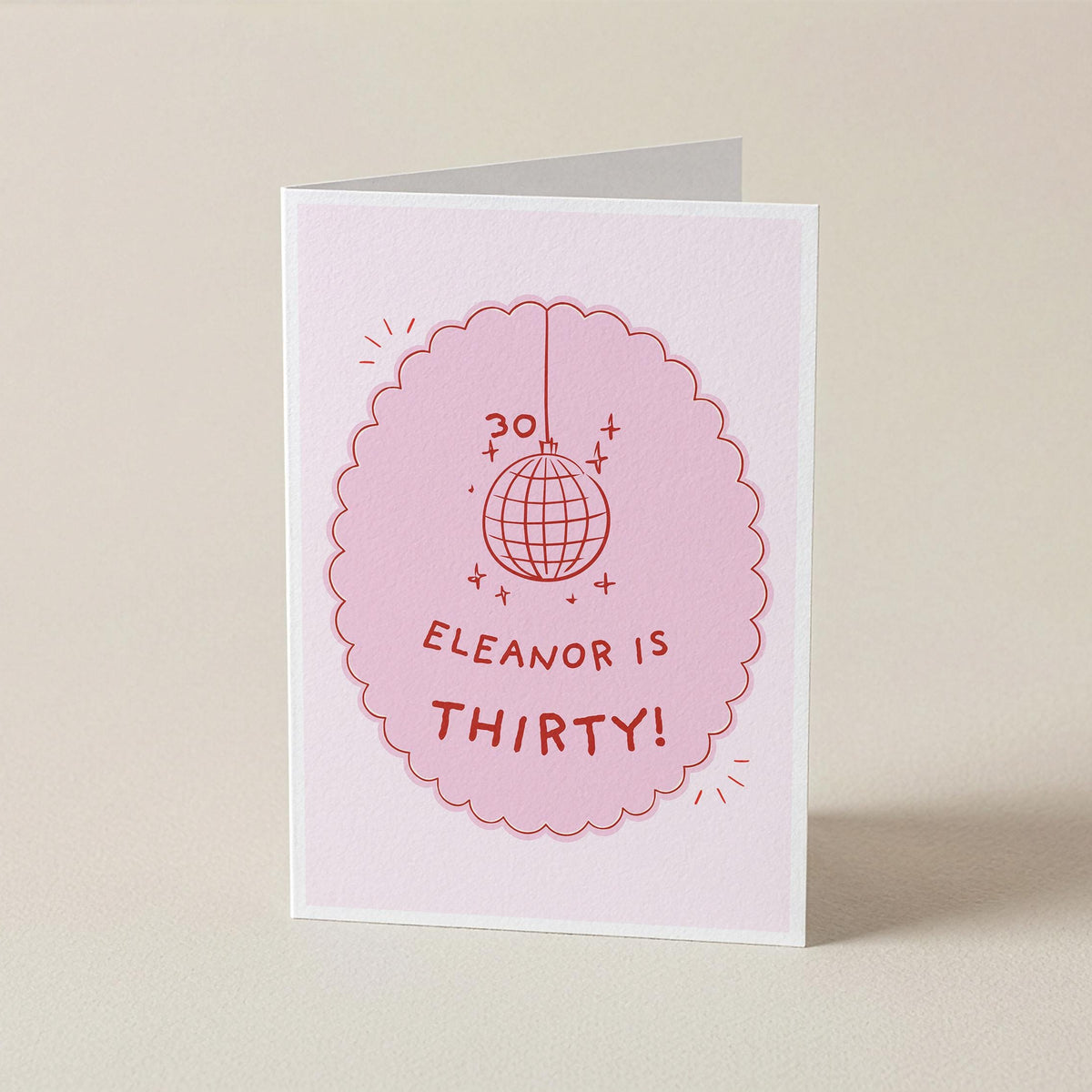 A folded greeting card standing upright on a light beige surface. The card is white with a pink scalloped oval graphic featuring a disco ball illustration and the text 'ELEANOR IS THIRTY!'.