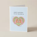 A folded greeting card standing upright on a light beige surface. The card has a pale blue background with the text 'HAPPY BIRTHDAY TO MY FAVOURITE' above a pink heart illustration featuring two cartoon Gemini characters and the word 'GEMINI'.