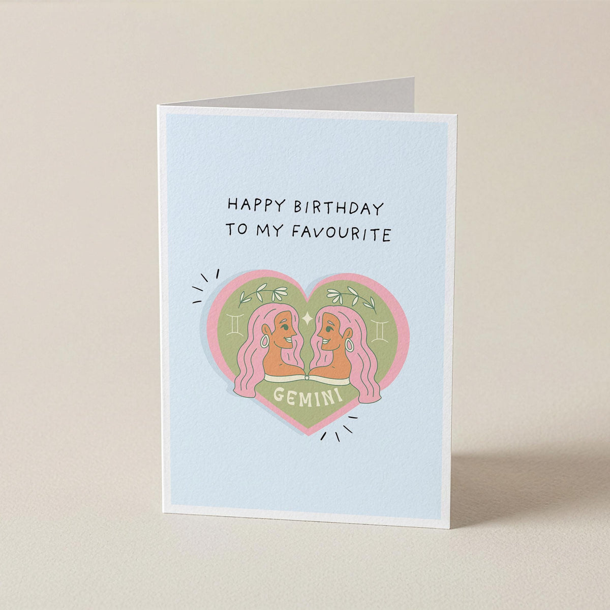 A folded greeting card standing upright on a light beige surface. The card has a pale blue background with the text 'HAPPY BIRTHDAY TO MY FAVOURITE' above a pink heart illustration featuring two cartoon Gemini characters and the word 'GEMINI'.