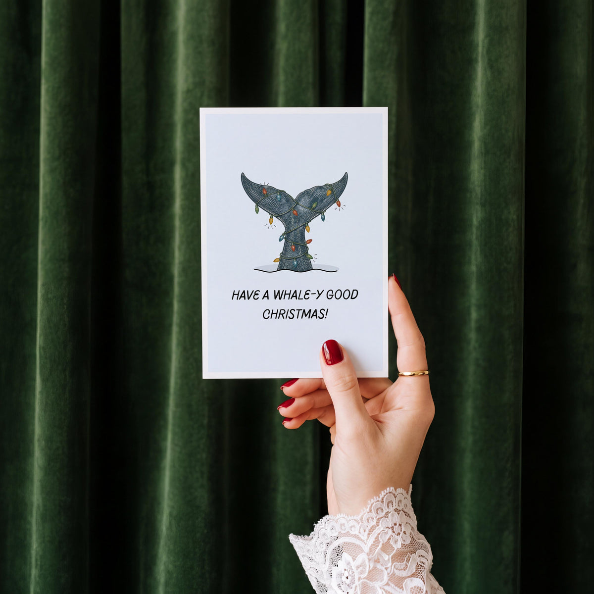 A hand with red nail polish and a white lace cuff holds a white greeting card featuring a mermaid tail illustration and the text 'HAVE A WHALE-Y GOOD CHRISTMAS!' against a dark green velvet curtain background.