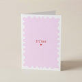 A folded greeting card with a white border and a light pink scalloped center, featuring the word 'SISTER' in red text above a small red heart, standing on a beige background.