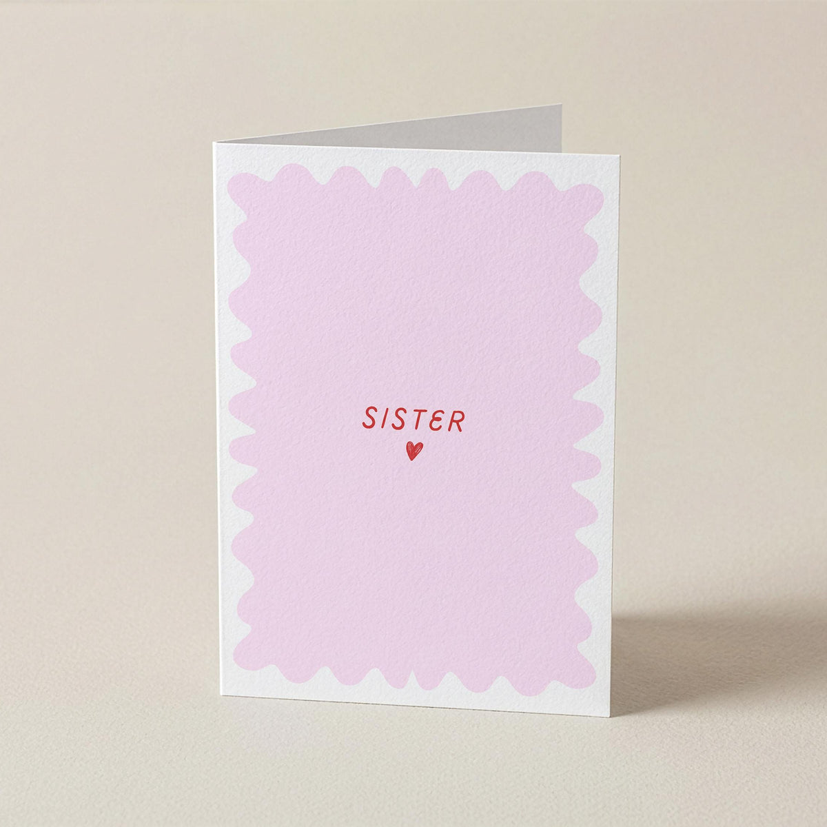 A folded greeting card with a white border and a light pink scalloped center, featuring the word 'SISTER' in red text above a small red heart, standing on a beige background.