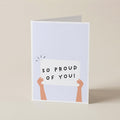 A folded greeting card standing upright on a light beige surface. The card has a pale blue front with a simple illustration of two peach-colored arms holding a white sign that reads “SO PROUD OF YOU!” in black uppercase letters.