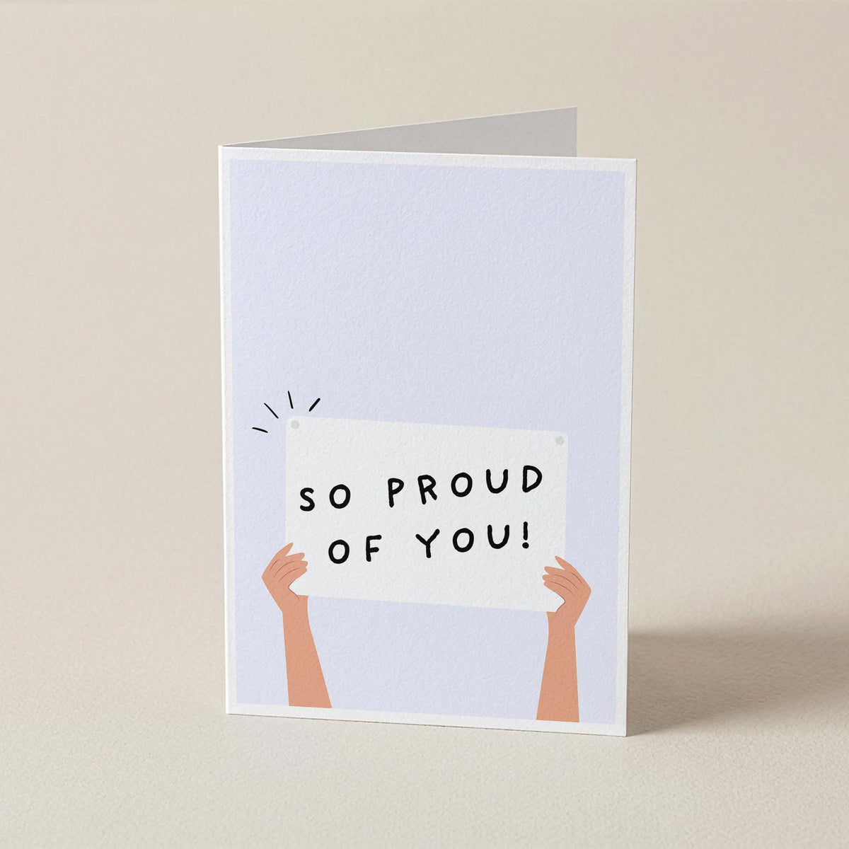 A folded greeting card standing upright on a light beige surface. The card has a pale blue front with a simple illustration of two peach-colored arms holding a white sign that reads “SO PROUD OF YOU!” in black uppercase letters.