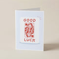 A folded greeting card standing upright on a light beige surface. The card has a pale blue border and features a white central panel with the words 'GOOD LUCK' in red above and below a red playing-card illustration.