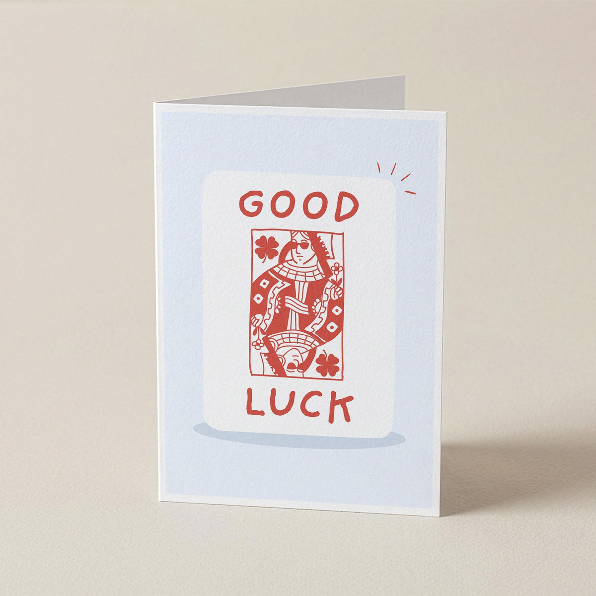 A folded greeting card standing upright on a light beige surface. The card has a pale blue border and features a white central panel with the words 'GOOD LUCK' in red above and below a red playing-card illustration.