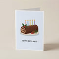 A folded greeting card standing upright on a light beige surface. The card features an illustration of a log cake with six lit candles, holly leaves, and pinecones, with the text 'HAPPY BIRTH-MAS!' printed below.