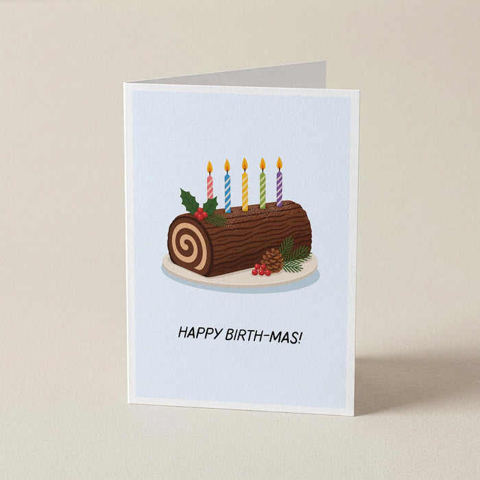 A folded greeting card standing upright on a light beige surface. The card features an illustration of a log cake with six lit candles, holly leaves, and pinecones, with the text 'HAPPY BIRTH-MAS!' printed below.