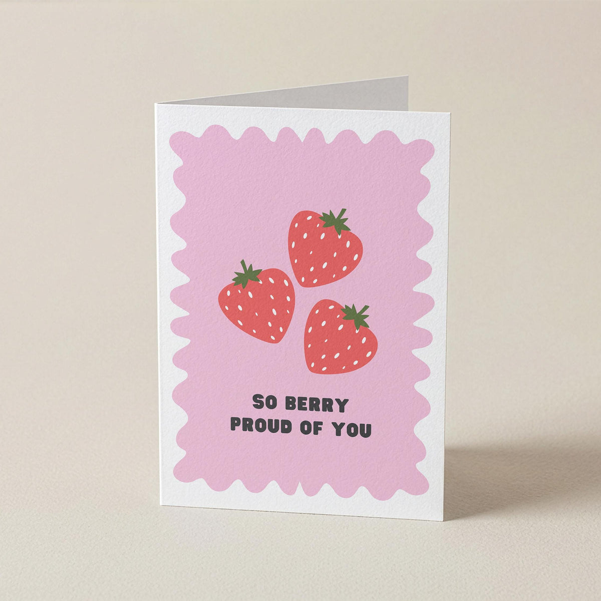 A folded greeting card standing upright on a light beige surface. The card has a pink scalloped border, three illustrated strawberries, and the text 'SO BERRY PROUD OF YOU' in black.