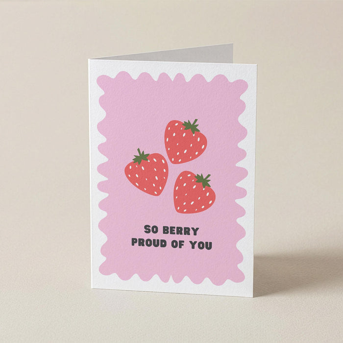 A folded greeting card standing upright on a light beige surface. The card has a pink scalloped border, three illustrated strawberries, and the text 'SO BERRY PROUD OF YOU' in black.