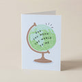 A folded greeting card standing upright on a beige surface. The card features an illustration of a globe with the text 'DAD YOU MEAN THE WORLD TO ME' in black lettering.