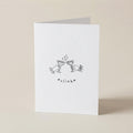 A white greeting card standing upright on a light beige surface, featuring a small black line drawing of two clinking glasses with the word '*clink*' beneath it.