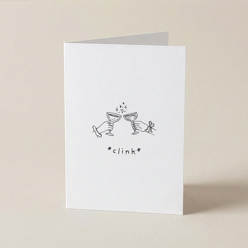 A white greeting card standing upright on a light beige surface, featuring a small black line drawing of two clinking glasses with the word '*clink*' beneath it.