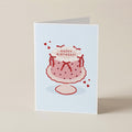 A folded greeting card standing upright on a light beige surface. The card has a pale blue front featuring a simple illustration of a pink frosted birthday cake with red polka dots, red bows, small red hearts, and the words “HAPPY BIRTHDAY!” on top.