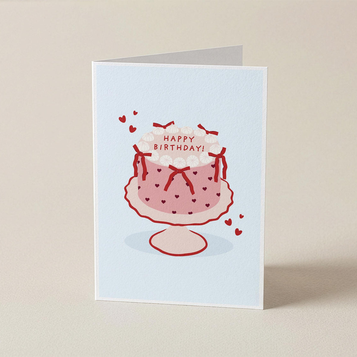 A folded greeting card standing upright on a light beige surface. The card has a pale blue front featuring a simple illustration of a pink frosted birthday cake with red polka dots, red bows, small red hearts, and the words “HAPPY BIRTHDAY!” on top.