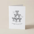 A white greeting card standing upright on a light beige surface, featuring a black line drawing of stacked party hats with the word 'congratulations!' below.