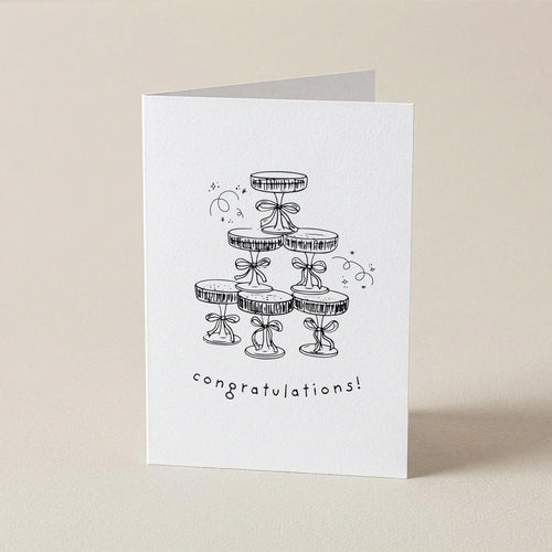 A white greeting card standing upright on a light beige surface, featuring a black line drawing of stacked party hats with the word 'congratulations!' below.