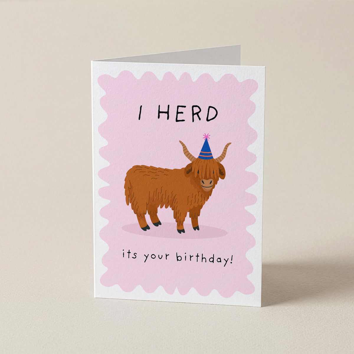 A folded greeting card standing upright on a light beige surface. The card has a pink scalloped border and features an illustration of a brown highland cow wearing a blue party hat, with the text 'I HERD it's your birthday!'.