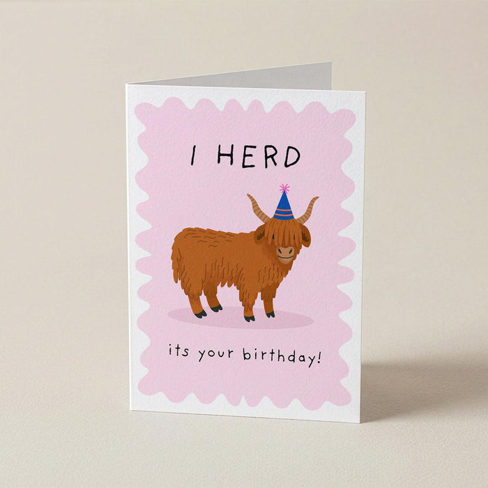 A folded greeting card standing upright on a light beige surface. The card has a pink scalloped border and features an illustration of a brown highland cow wearing a blue party hat, with the text 'I HERD it's your birthday!'.