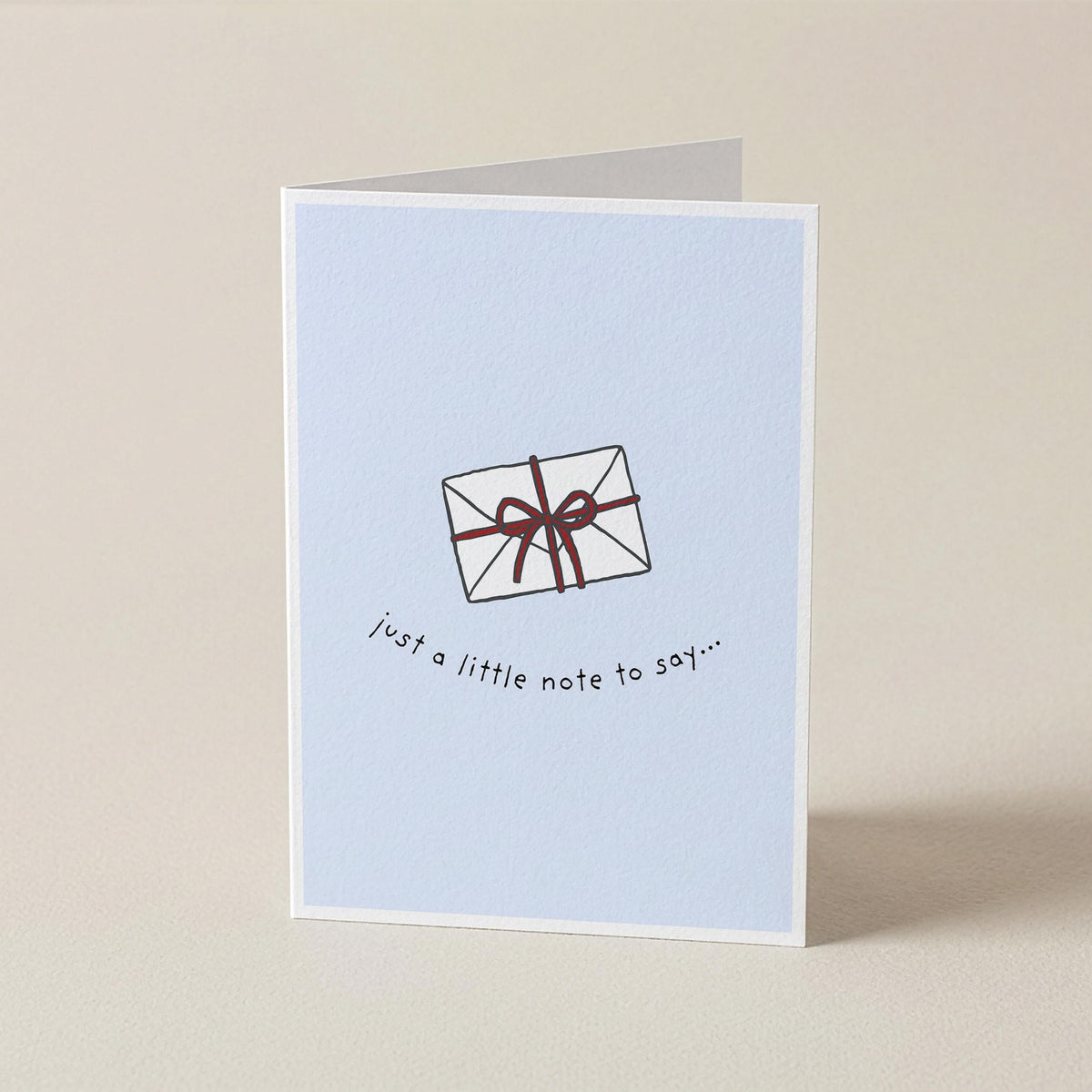 A folded greeting card standing upright on a light beige surface. The card has a pale blue front with a simple line drawing of a wrapped gift and the text 'just a little note to say.