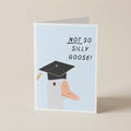 A folded greeting card standing upright on a light beige surface. The card has a pale blue front with the text 'NOT SO SILLY GOOSE!' in black and an illustration of a goose wearing a black graduation cap.