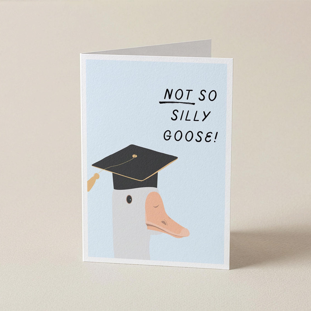 A folded greeting card standing upright on a light beige surface. The card has a pale blue front with the text 'NOT SO SILLY GOOSE!' in black and an illustration of a goose wearing a black graduation cap.