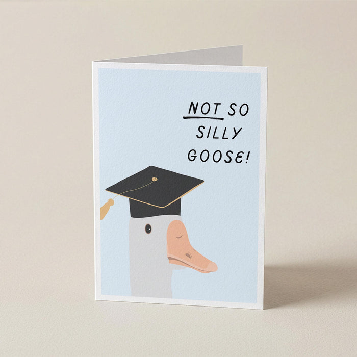 A folded greeting card standing upright on a light beige surface. The card has a pale blue front with the text 'NOT SO SILLY GOOSE!' in black and an illustration of a goose wearing a black graduation cap.