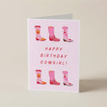 A folded greeting card standing upright on a light beige surface. The card is pale pink with the text 'HAPPY BIRTHDAY COWGIRL!' in red and illustrations of pink cowboy boots with red hearts and flames.
