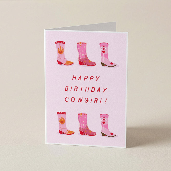 A folded greeting card standing upright on a light beige surface. The card is pale pink with the text 'HAPPY BIRTHDAY COWGIRL!' in red and illustrations of pink cowboy boots with red hearts and flames.