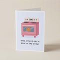 A folded greeting card standing upright on a light beige surface. The card features a minimalist illustration of a pink oven with the text 'OMG, YOU'VE GOT A BUN IN THE OVEN!' printed below it.