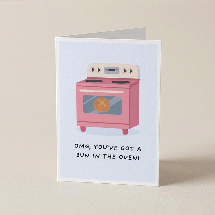 A folded greeting card standing upright on a light beige surface. The card features a minimalist illustration of a pink oven with the text 'OMG, YOU'VE GOT A BUN IN THE OVEN!' printed below it.