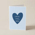 A folded greeting card standing upright on a light beige surface. The card has a pale blue front with a large dark blue heart in the center containing the white text 'LOVE YOU, BRO'.