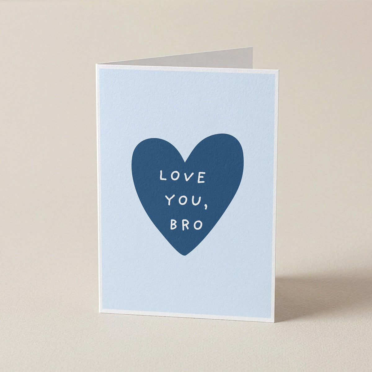 A folded greeting card standing upright on a light beige surface. The card has a pale blue front with a large dark blue heart in the center containing the white text 'LOVE YOU, BRO'.