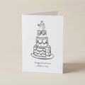 A white greeting card standing upright on a light beige surface, printed with a black line drawing of a three-tier wedding cake and the text 'Congratulations Jake & Lily' beneath it.