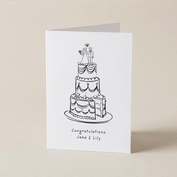 A white greeting card standing upright on a light beige surface, printed with a black line drawing of a three-tier wedding cake and the text 'Congratulations Jake & Lily' beneath it.