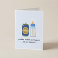 A folded greeting card standing upright on a light beige surface. The card has a white front with a simple illustration of a blue baby bottle and a blue beer can with gold labels, plus the text 'HAPPY FIRST BIRTHDAY AS MY DADDY!'.